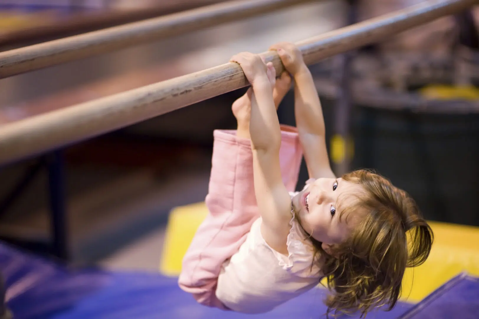 Young girl, aged 2, proving it is never to early to start having fun and practicing good physical fitness. She is pulling herself up on uneven bars. Blue and yellow mats defocused in the background. Shallow DOF provides good subject isolation, focus on her eyes.